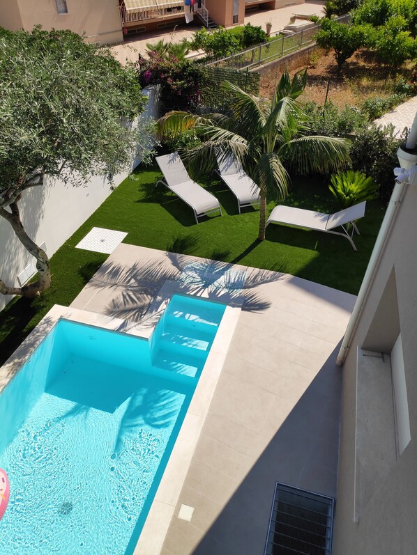 Ein Schwimmbecken mit gefliestem Deck, zwei Liegestühlen und einer kleinen Gartenanlage mit einer Palme.