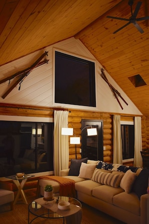 Ein Holzblockhaus-Innenraum mit hoher Decke, einem großen Fenster, einer Couch, einem Couchtisch und einer Lampe.