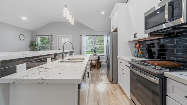 Une cuisine moderne avec des meubles blancs, un grand îlot central, des appareils électroménagers en acier inoxydable et une vue sur l’extérieur.