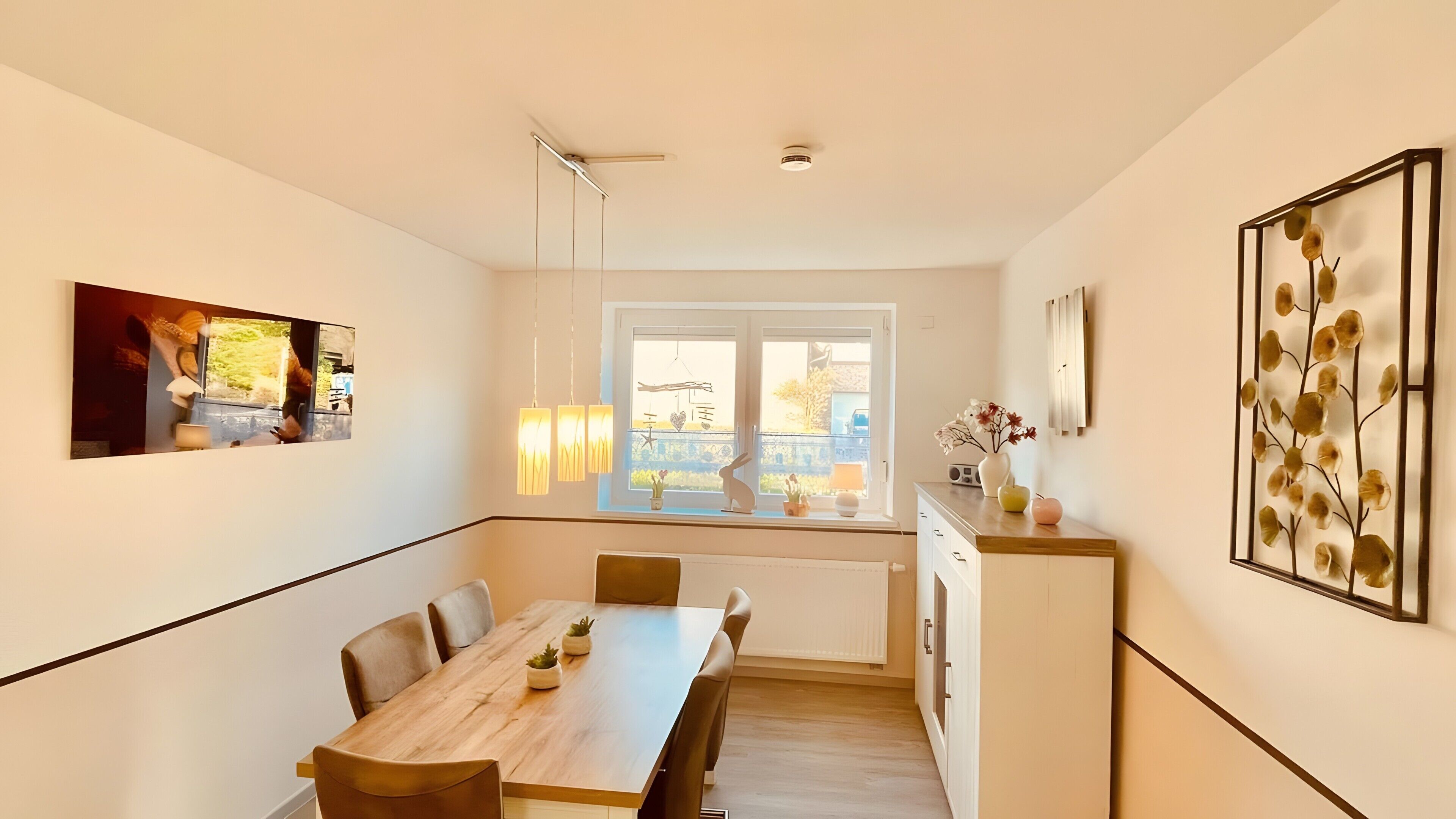 A modern kitchen with a dining area, a large window with a view, pendant lights, and a decorative wall piece.