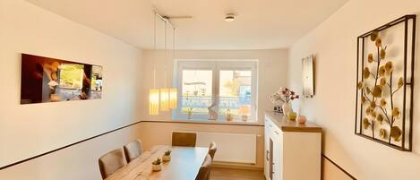 A modern kitchen with a dining area, a large window with a view, pendant lights, and a decorative wall piece.