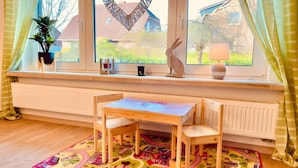 Ein Zimmer mit Fensterbank, Tisch, Stühlen, Heizkörper, Topfpflanze und einem farbenfrohen Teppich.
