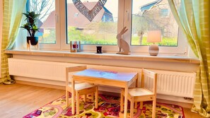 Ein Zimmer mit Fensterbank, Tisch, Stühlen, Heizkörper, Topfpflanze und einem farbenfrohen Teppich.