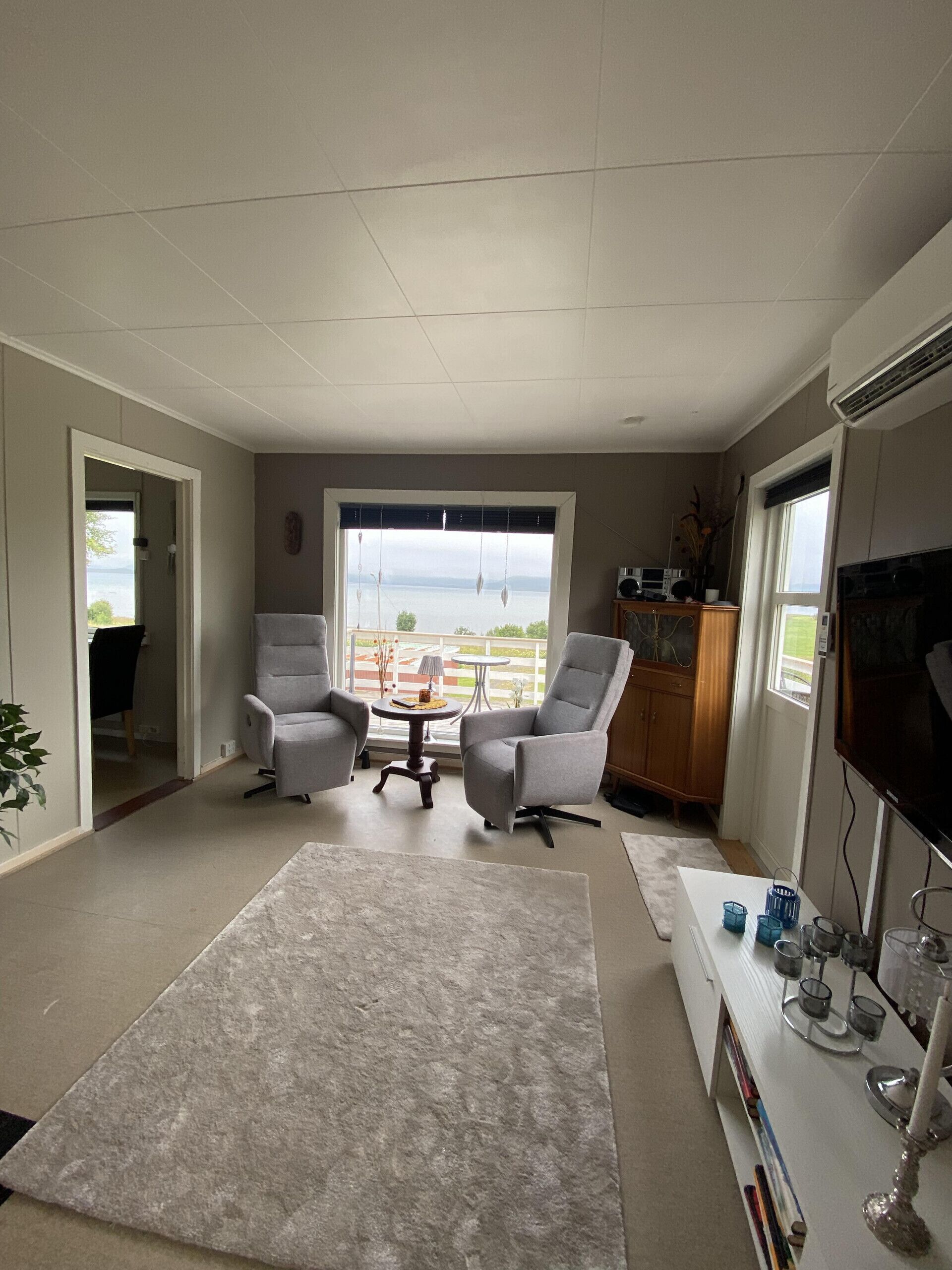 Ein modernes Wohnzimmer mit einem großen Fenster, zwei grauen Sesseln, einem weißen Couchtisch und einem Flachbildfernseher.