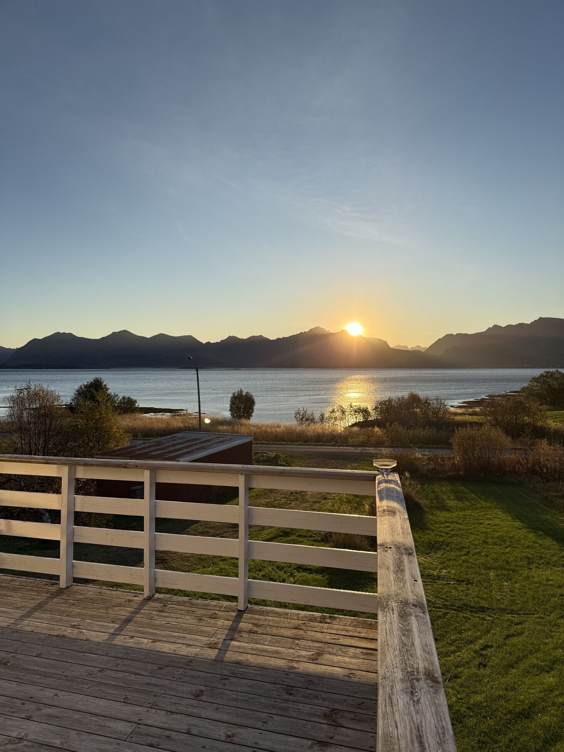 Una terraza de madera con baranda, vista a un cuerpo de agua con montañas al fondo y el sol poniéndose.