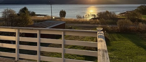 A wooden deck with a railing, overlooking a body of water with mountains in the distance and the sun setting.