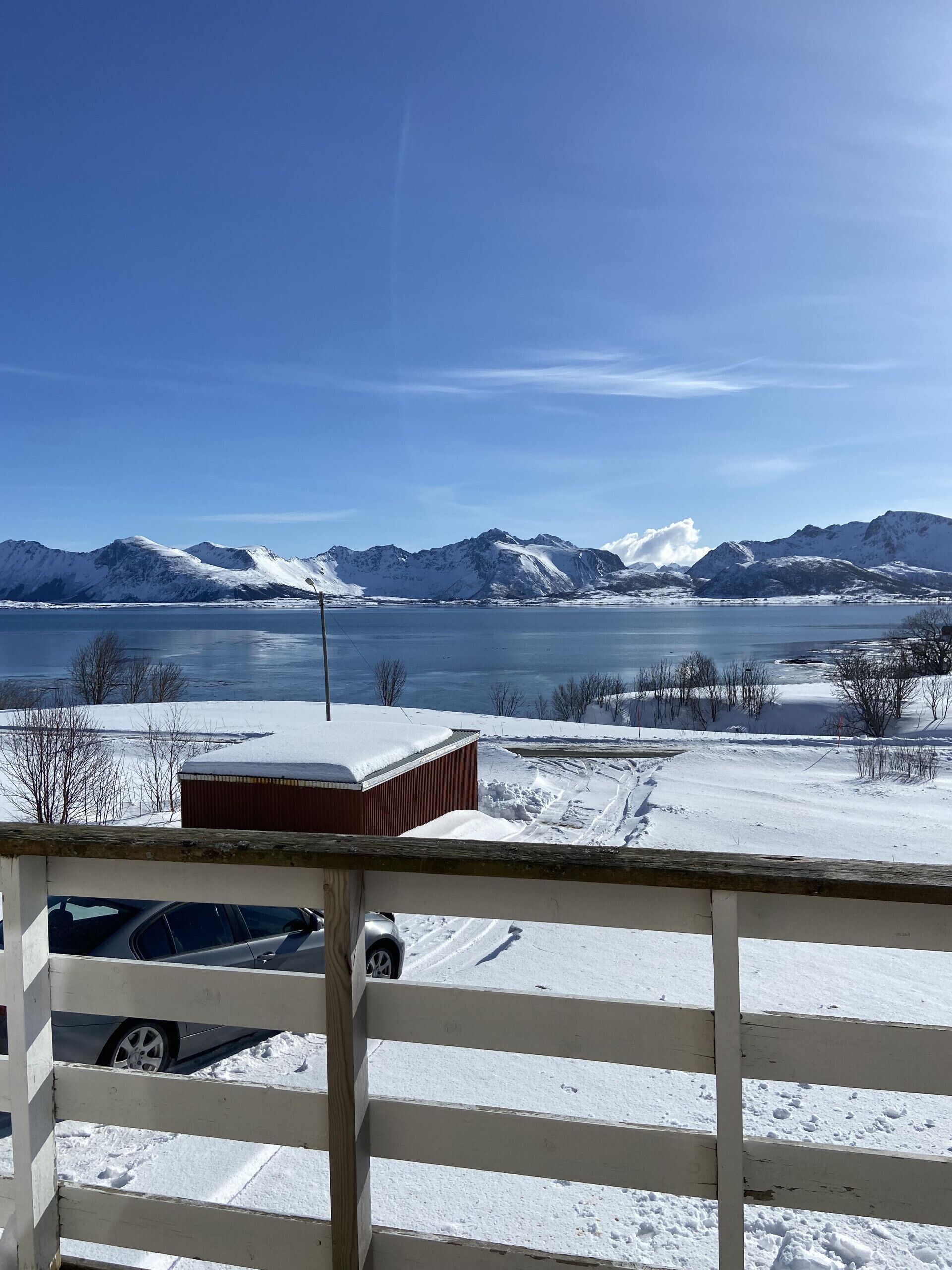 A snowy landscape with mountains in the background, a wooden fence, and a building with snow on its roof.
