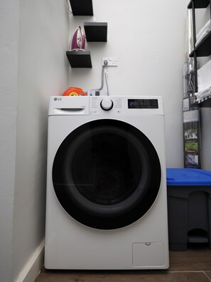 Una lavadora LG de carga frontal en un cuarto de lavado, con un tapete azul, un estante con productos de limpieza y un enchufe.