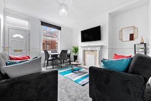 A modern living room with a fireplace, a flat-screen TV, and a dining area. - Spacious Leeds Home | Sleeps 6 | Free Parking (Beeston)