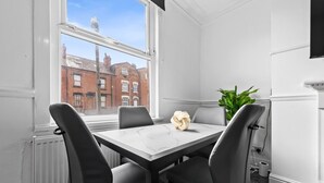 Quarto com uma mesa branca, quatro cadeiras, uma planta e uma janela com vista para edifícios.