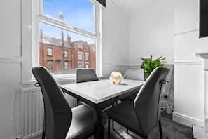 A room with a white table, four chairs, a plant, and a window with a view of buildings. - Spacious Leeds Home | Sleeps 6 | Free Parking (Beeston)
