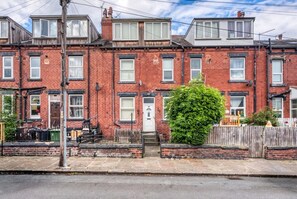 A row of red brick terraced houses with white windows, a green bush, and a wooden fence. - Spacious Leeds Home | Sleeps 6 | Free Parking (Beeston)