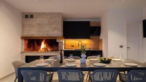 A modern dining area with a fireplace, a table set for six, and a kitchen in the background. - Villa Freedom (Čista Velika)