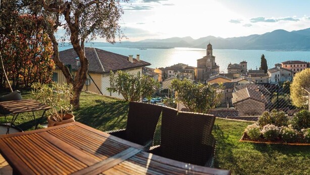 Vista de un pueblo a orillas de un lago, con edificios, una iglesia y un área de comedor con mobiliario exterior.