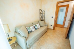 A room with a beige sofa, a small round table with a lamp, a decorative metal piece on the wall, and a wooden door. - Apartment 'Jessica' with Lake View, Wi-Fi and Air Conditioning (Torri del Benaco)