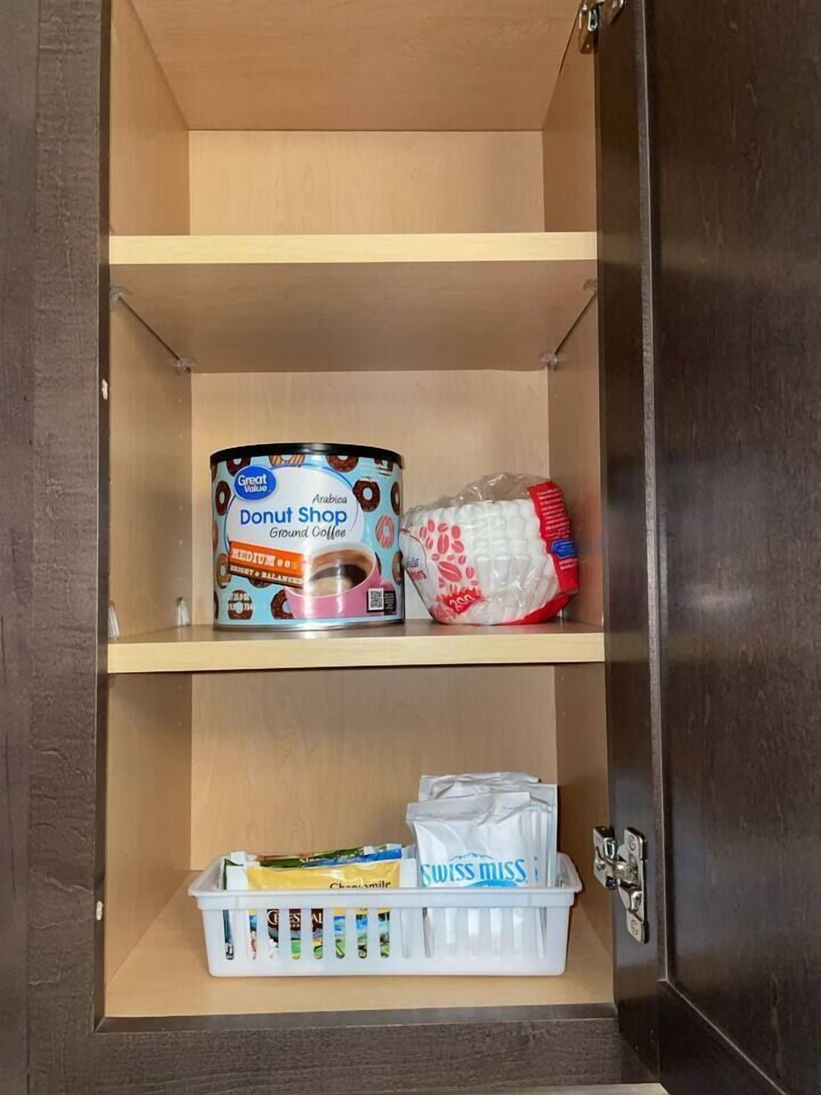Ein Schrank mit einer Dose Great Value Arabica Donut Shop Ground Coffee Medium auf der obersten Ablage, einer Plastiktüte mit rotem und weißem Design auf der mittleren Ablage und einem weißen Korb mit verschiedenen Artikeln auf der untersten Ablage.