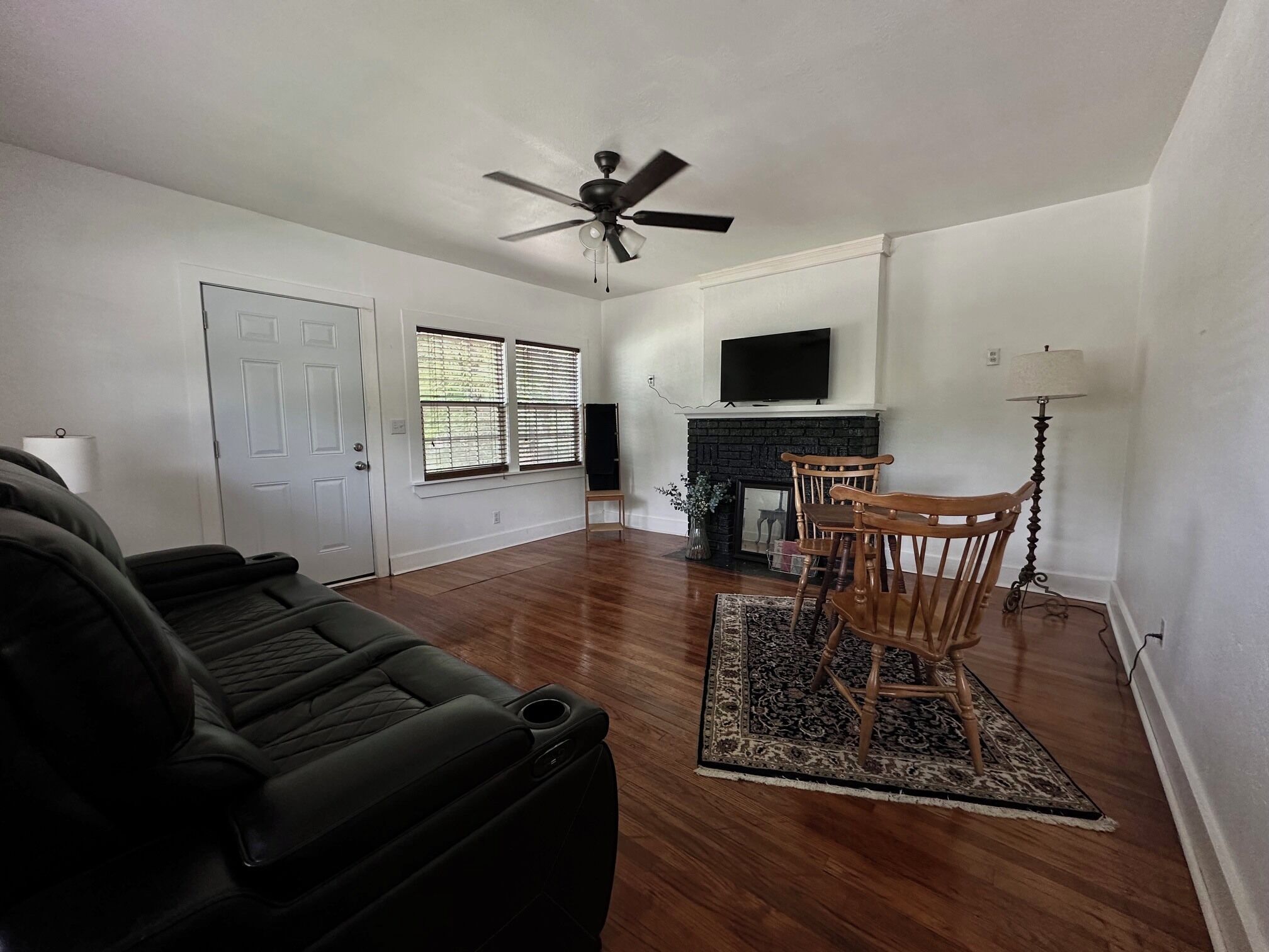 A living room with a black leather couch, a wooden dining table with chairs, a fireplace, and a ceiling fan.