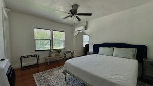 Quarto com cama, ventoinha no teto, escrivaninha com cadeira, janela com persianas e radiador.