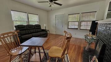 A living room with a sofa, two wooden chairs, a table, a ceiling fan, and a fireplace.