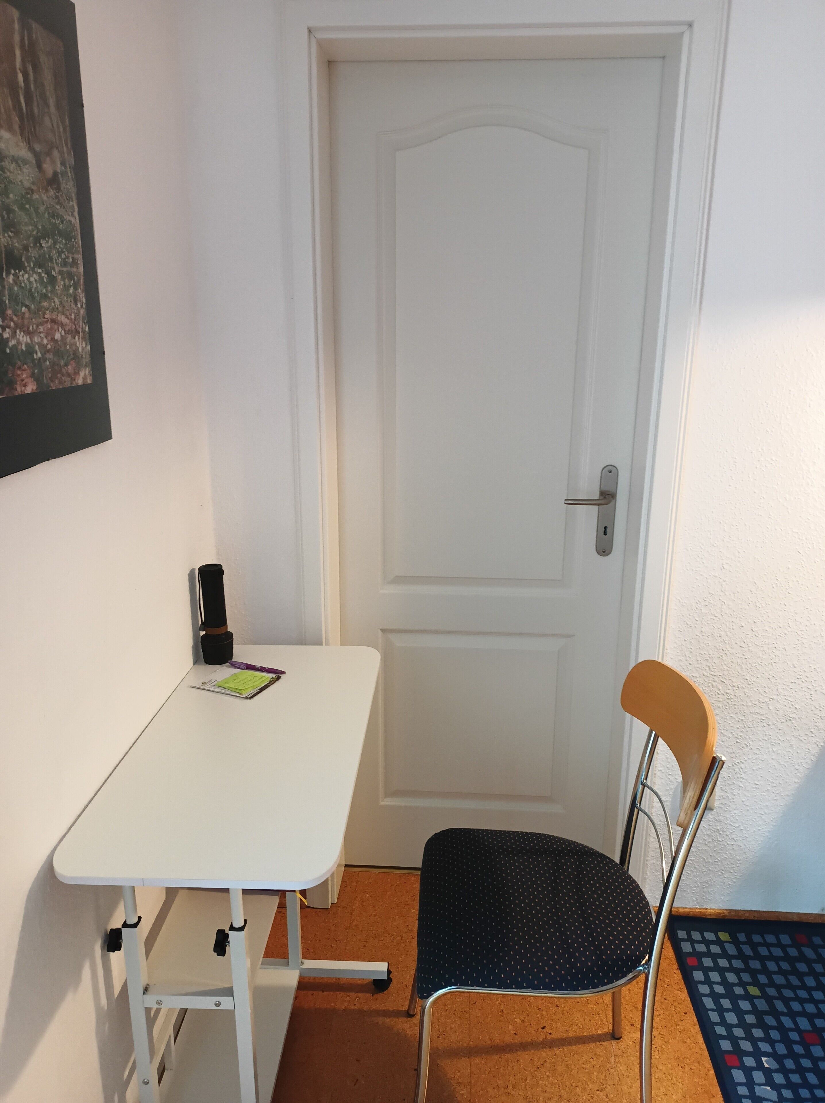Ein kleiner Raum mit einer weißen Tür, einem weißen Tisch mit Notizbuch und Stift, einem schwarzen Stuhl mit Holzlehne und einem bunten Teppich auf dem Boden.