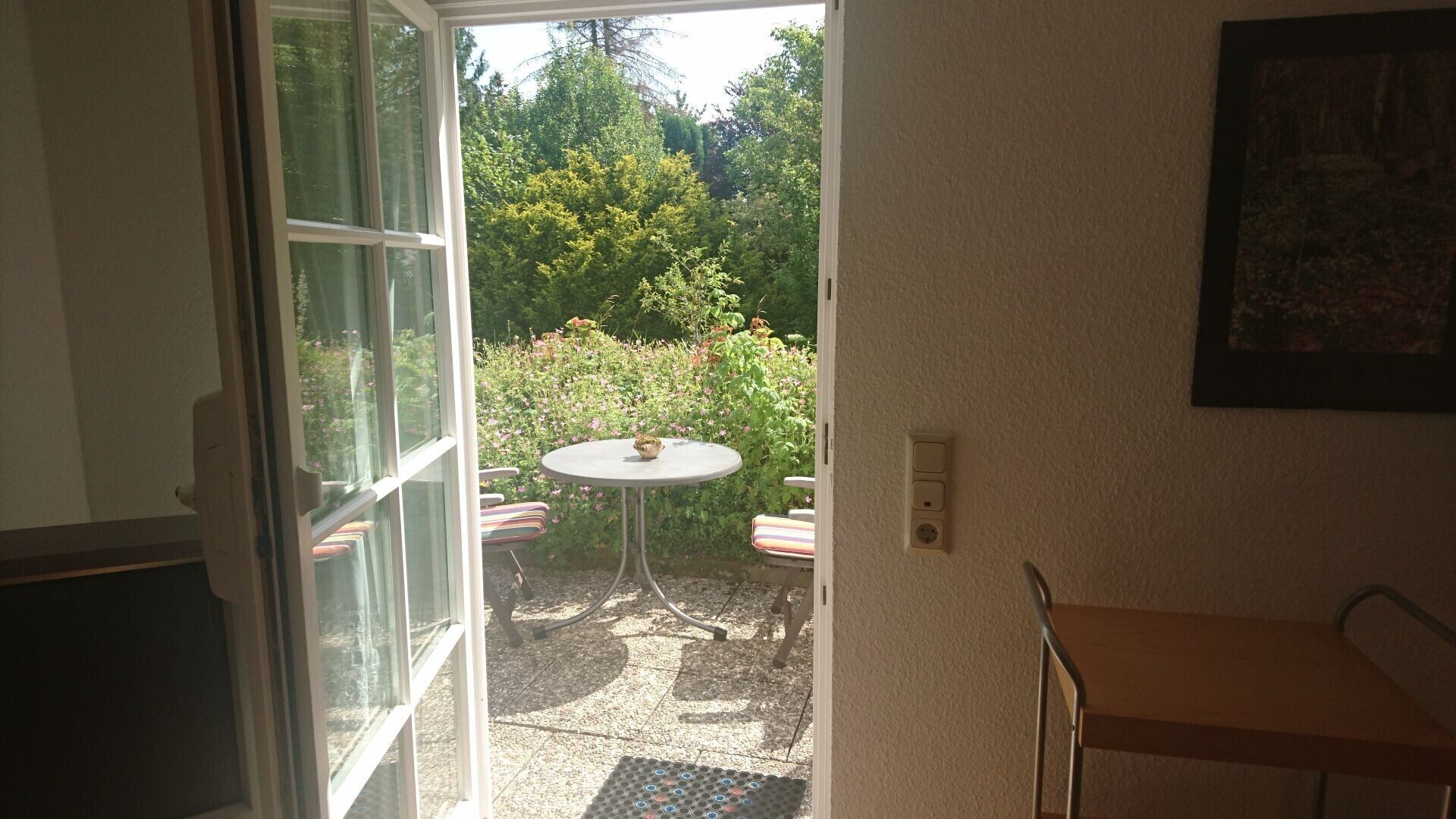 Une porte ouverte donnant sur une terrasse avec une table et des chaises, un petit jardin et une prise de courant sur le mur.