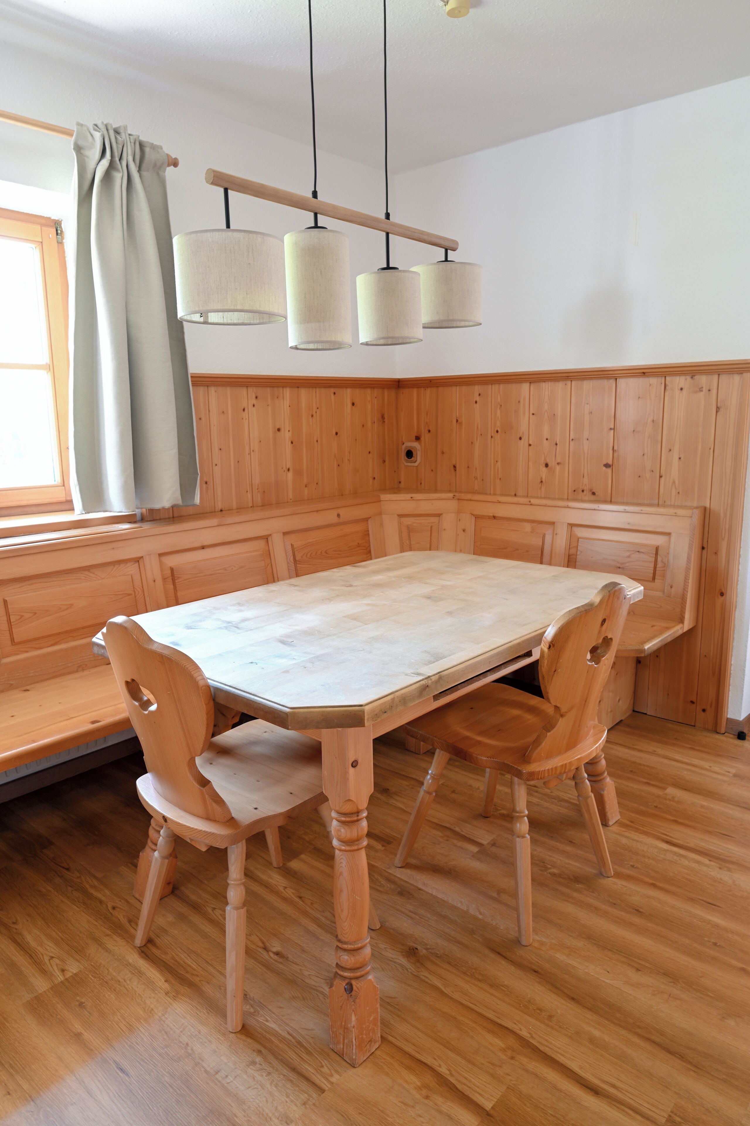 A wooden dining table with bench seating, a hanging light fixture, and a window with curtains.