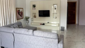 Una sala de estar moderna con un sofá gris, un televisor de pantalla plana montado en una pared blanca, una pequeña mesita con una planta, y una puerta que da a otra habitación.
