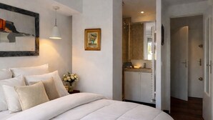 Een slaapkamer met een groot bed, een nachtkastje met een vaas, een lijst met een afbeelding en een gang die naar een badkamer leidt.