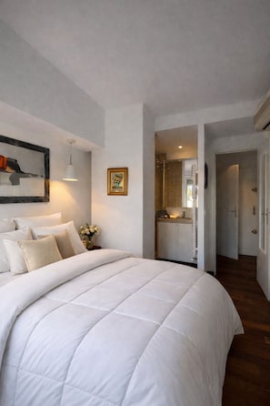A bedroom with a large bed, a bedside table with a vase, a framed picture, and a hallway leading to a bathroom. - Vercors Festivals next to the sea & beach (Cannes)