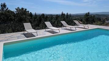 En rektangulär pool med klart, blått vatten, omgiven av solstolar och en stenbelagd terrass.