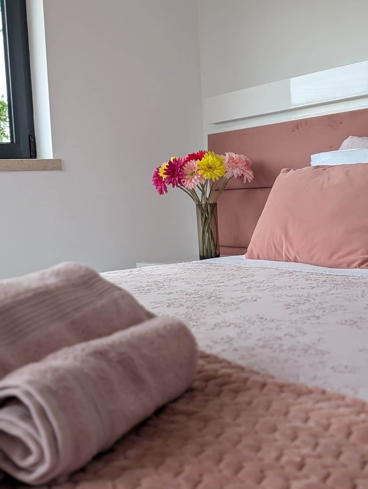 Una cama bien hecha con una toalla enrollada, un cabecero rosa y un jarrón con flores sobre la mesita de noche.