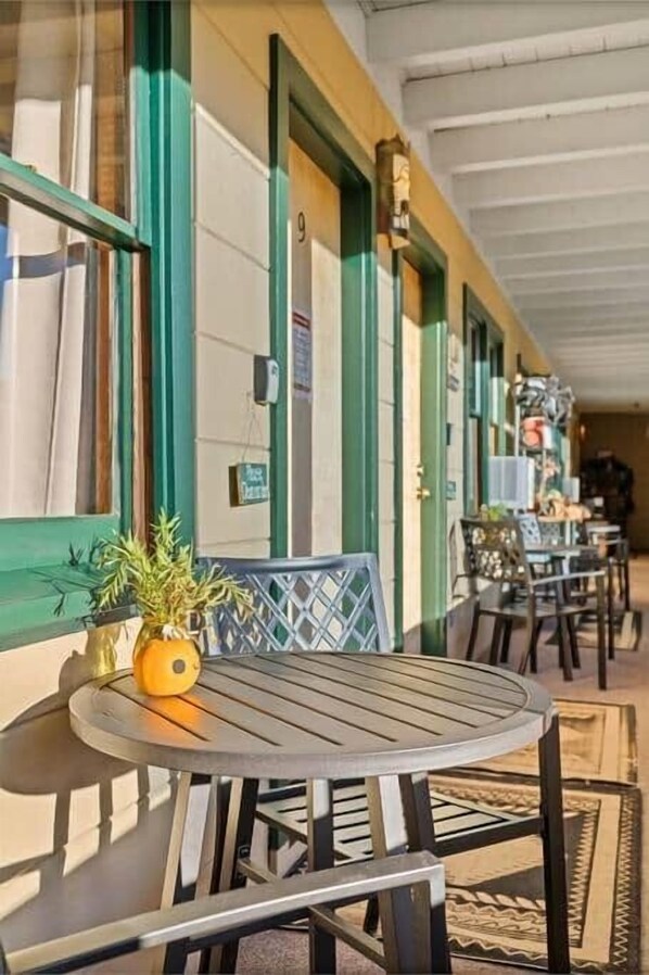 A covered outdoor seating area with tables and chairs, a potted plant, and a building with green trim and numbered doors.