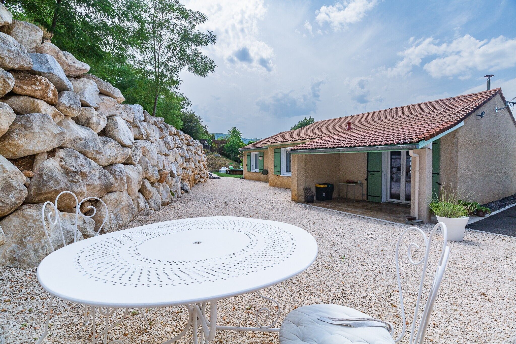 A stone wall, a small house with a tiled roof, and a round table with chairs in a gravel area.