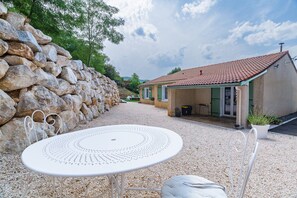 A stone wall, a small house with a tiled roof, and a round table with chairs in a gravel area. - Holiday Home 'Le Beauregard - Piscine' with Mountain View, Wi-Fi and Air Conditioning (Beauregard-Baret)