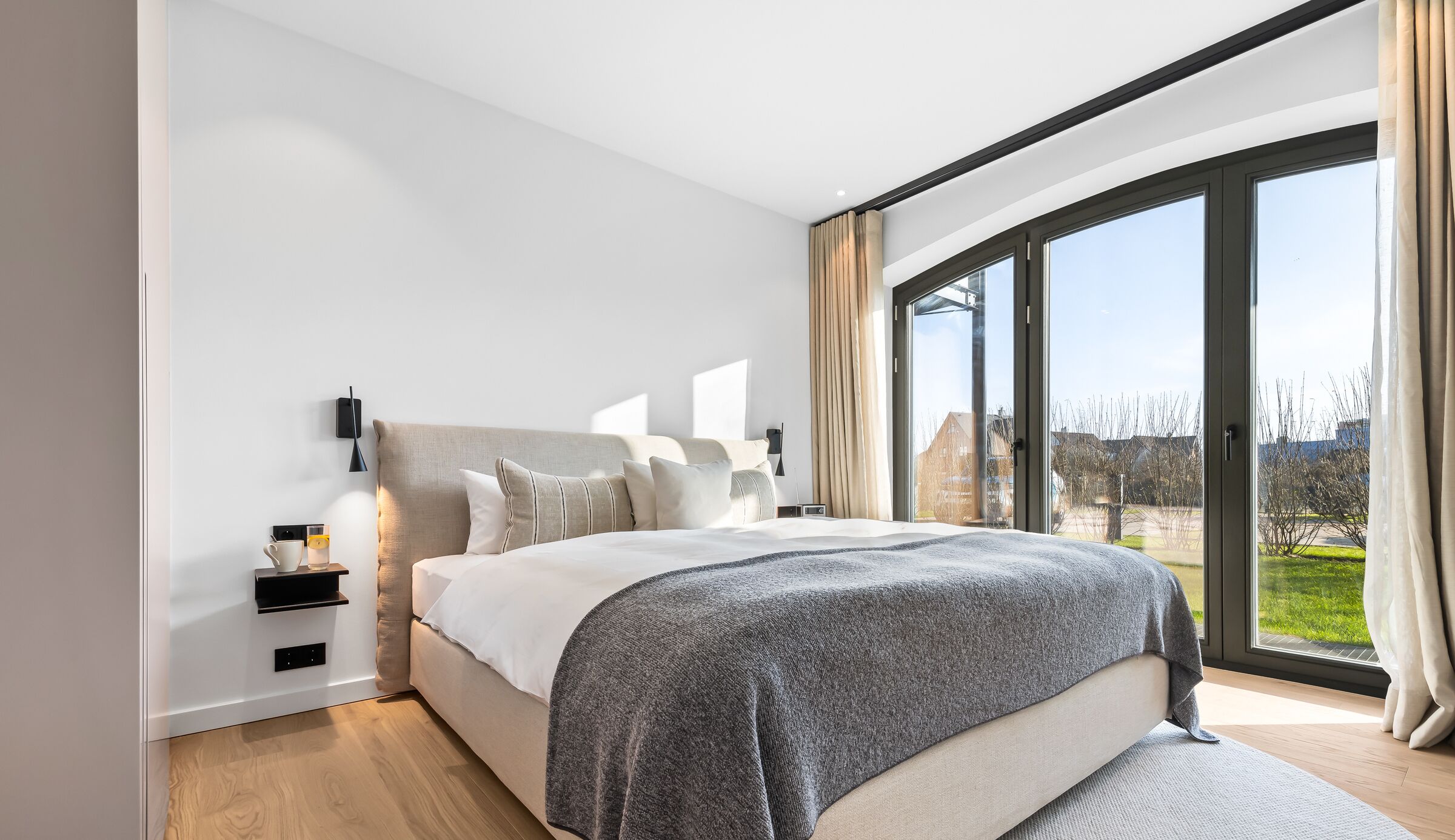 A modern bedroom with a large bed, a nightstand, a wall-mounted lamp, and a view of the outdoors through a large window.