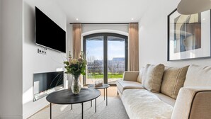 A modern living room with a flat-screen TV, a sofa, a coffee table, and a large window with curtains. - Hudson Suite, a luxurious  featuring both a Finnish sauna and an infrared sauna. (List auf Sylt)