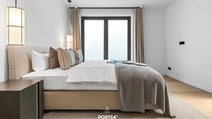 A modern bedroom with a bed, bedside tables, a hanging light fixture, and a large window with curtains. - Hudson Suite, a luxurious  featuring both a Finnish sauna and an infrared sauna. (List auf Sylt)