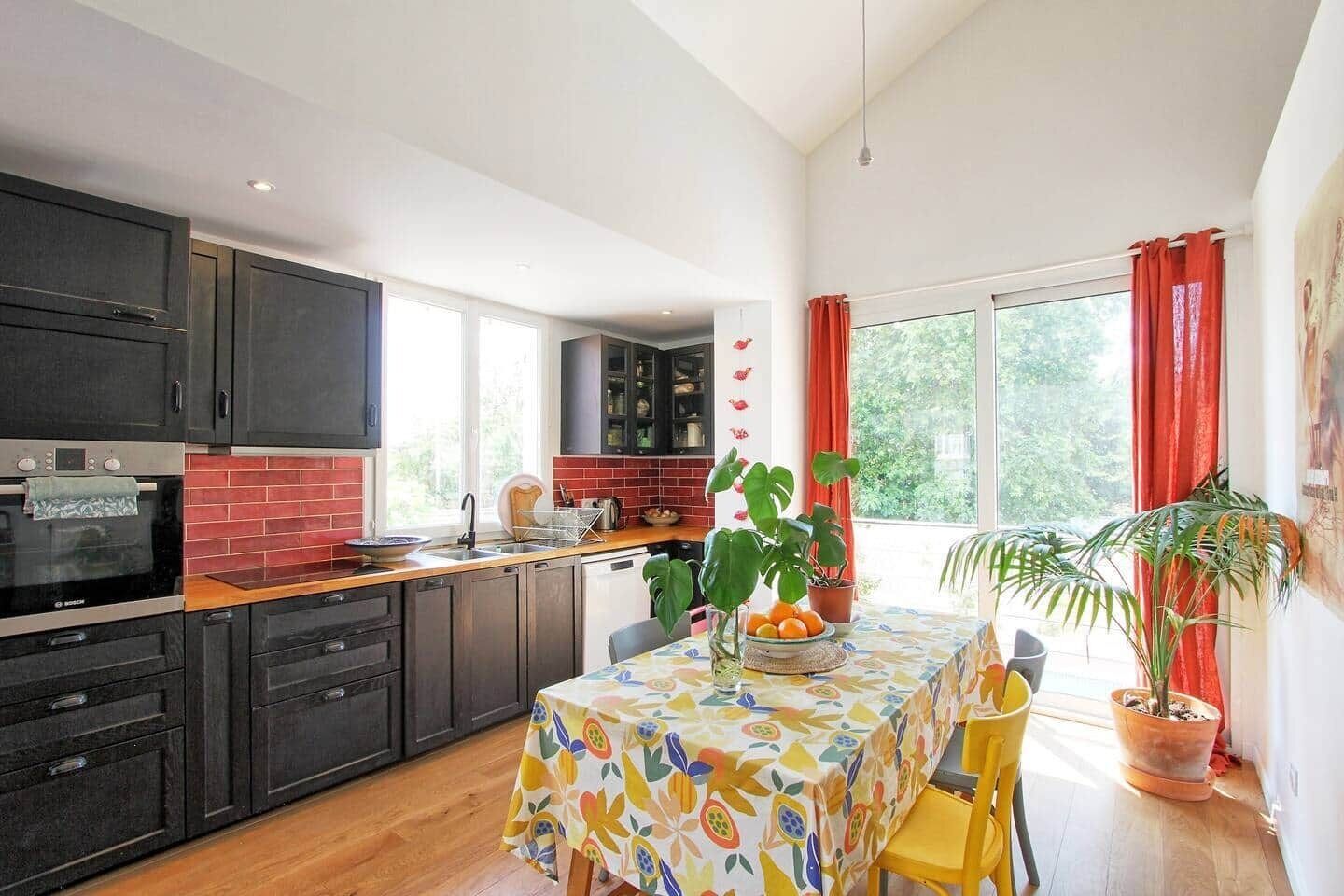 A kitchen with dark cabinets, a red brick backsplash, and a dining area with a colorful tablecloth.