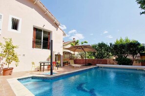 Une piscine avec un bord carrelé, une terrasse avec des chaises et une table, et un bâtiment doté d’une fenêtre et d’une porte.