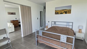 Una camera da letto con una struttura letto in metallo, un comodino bianco con una lampada, una sedia e una porta che conduce in un'altra stanza.