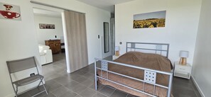 Et soveværelse med en seng med metalramme, et hvidt natbord med lampe, en stol og en dør, der fører til et andet rum.