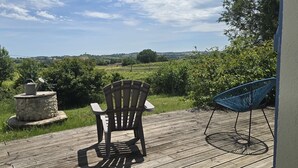Un patio in legno con una sedia e una poltrona blu, un vaso in pietra con un annaffiatoio, e vista su un paesaggio verde.