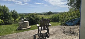 En træterrasse med en stol og en blå stol, en plantekasse i sten med en vandkande, samt udsigt over et grønt landskab.