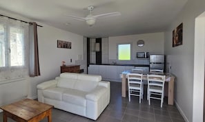 Un espai habitable compacte amb zona de cuina, un sofà blanc, una taula de fusta i un ventilador de sostre.