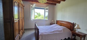 A bedroom with a wooden bed, a nightstand, a lamp, a wardrobe, and a window with a view of greenery.