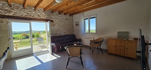 Habitación con pared de piedra, techo de madera, sofá, mesa, sillas, aparador de madera y vista a un jardín a través de una puerta de vidrio.