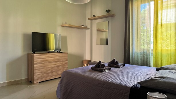 A modern bedroom with a bed, a TV on a wooden dresser, a bedside table, a mirror, and a window with curtains. - Apartment 2 rooms - house Martina LuxRental Apartments (Pescara)