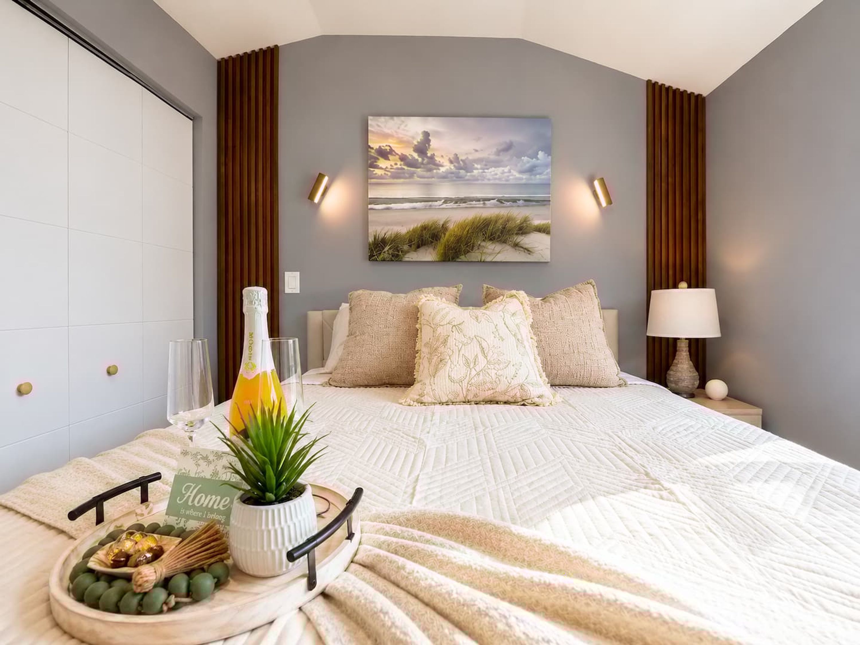 Un dormitorio con cama, mesita de noche con lámpara, un cuadro en la pared, y otra mesita de noche con una botella y una planta.