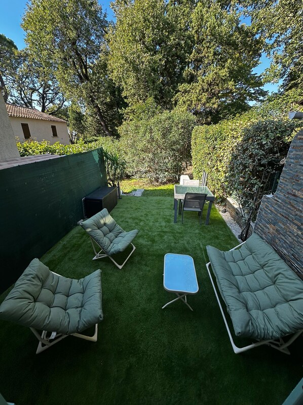 Un patio con tres sillones de descanso y una mesita, rodeado de césped artificial verde y árboles.