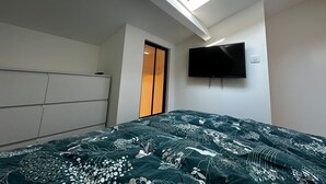 Quarto com claraboia, cama com colcha estampada, televisão na parede e guarda-roupa.